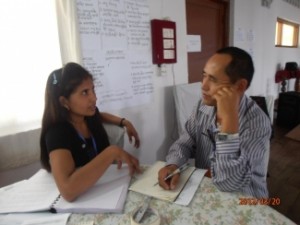 One female and one male monitor sit facing each other at a table. In front of them are books, papers and pens. They are close to a wall with flip chart notes taped to it.