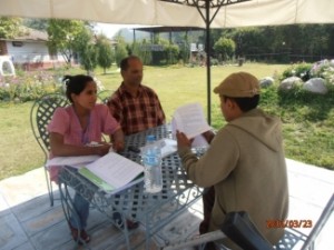 Three people, one woman and two men, are sitting outside, at a table covered by an umbrella. In the background, there is a gazebo to the right and another building to the left.